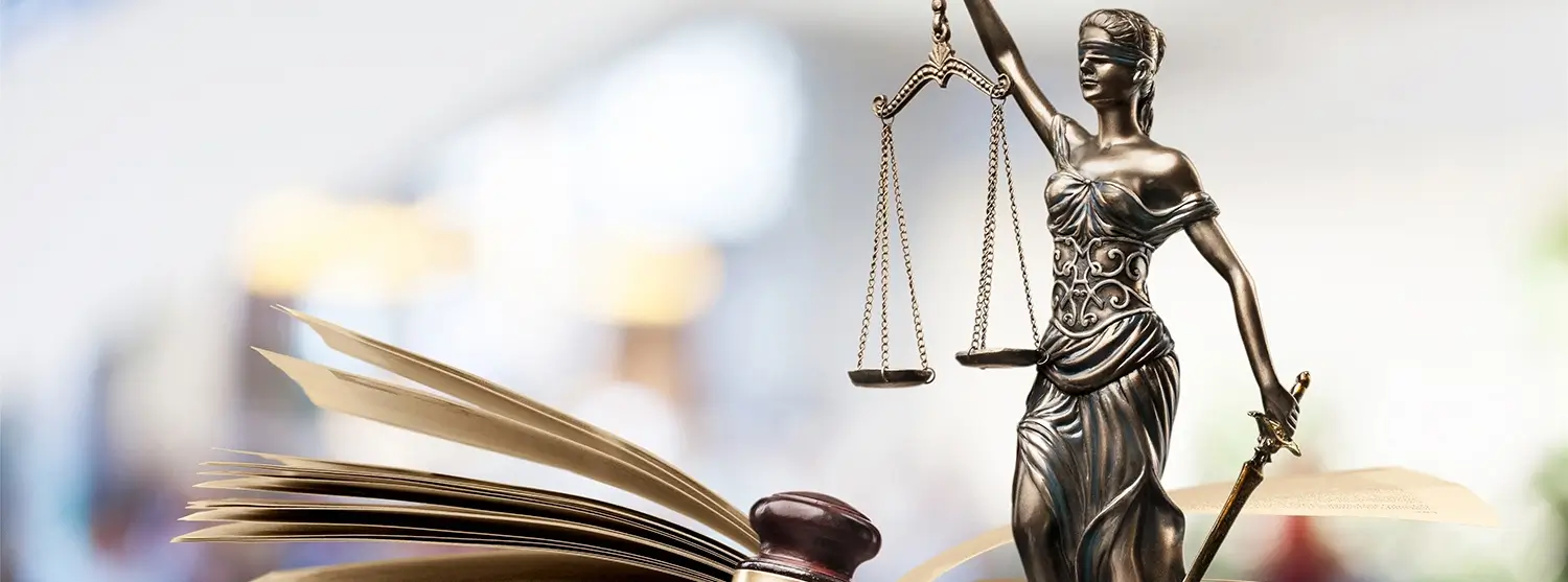 A bronze Lady Justice statue holding scales and a sword, standing next to an open law book, with a wooden gavel in the foreground.