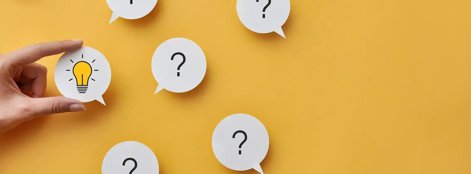 A yellow background with several white speech bubbles containing question marks, and one bubble showing a lightbulb, symbolizing an idea or solution. A hand is holding the bubble with the lightbulb.