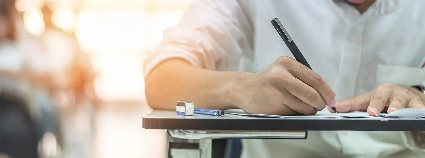 A close-up of a person in a white shirt writing with a pen on a paper at a desk, with a sharpener and pencil in the background.